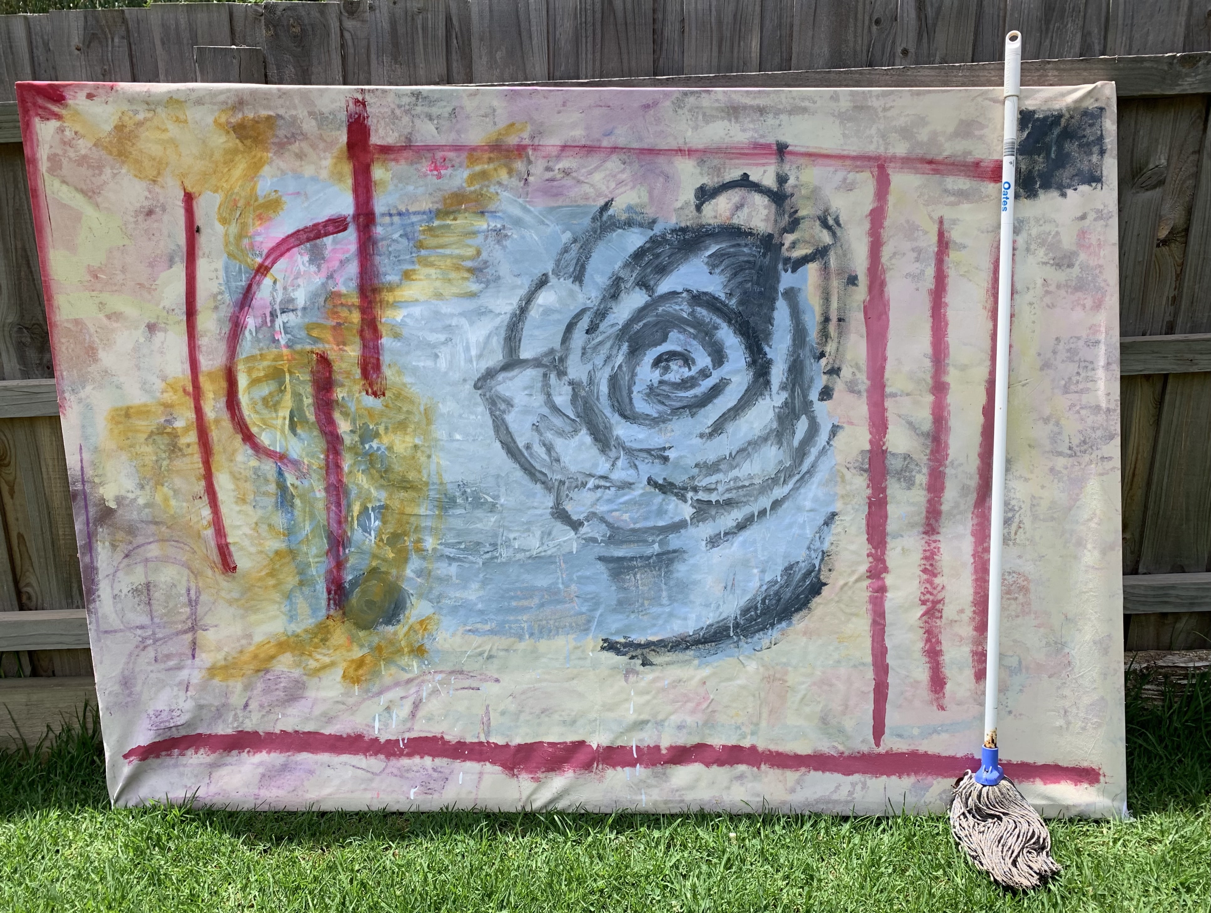A photograph of a canvas artwork. In situ to the artwork is a brown fence and bright green grass in the foreground. The canvas includes gestural movements, lines, brush marks and strong red markings. In the centre of the image is a blue shape and overlaid a rose drawn in navy paint.